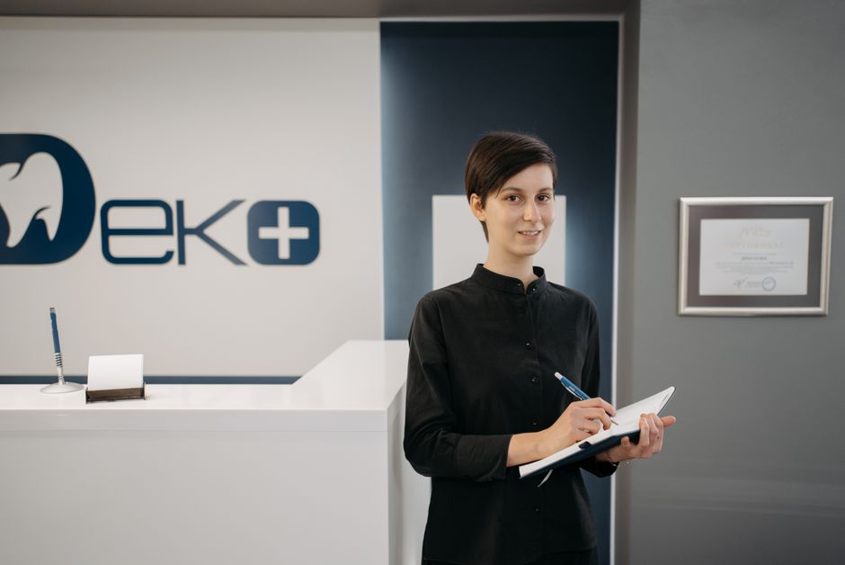 Pacjent przy okienku rejestracji w przychodni, przekazuje dokumenty pracownikowi. W tle poczekalnia z osobami czekającymi na 