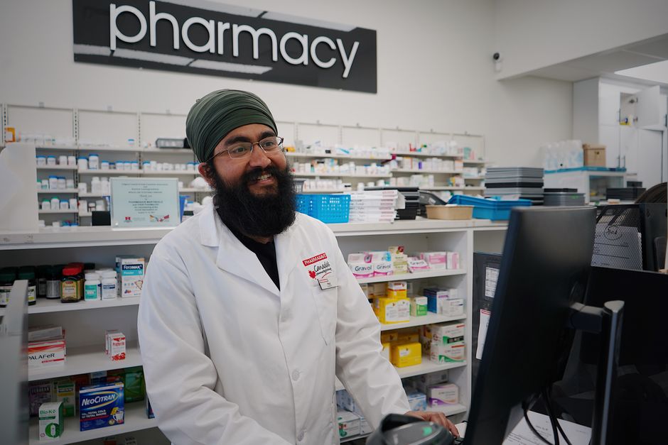 Farmaceutka w białym fartuchu podaje klientce opakowanie leku przy ladzie apteki z półkami pełnymi produktów.